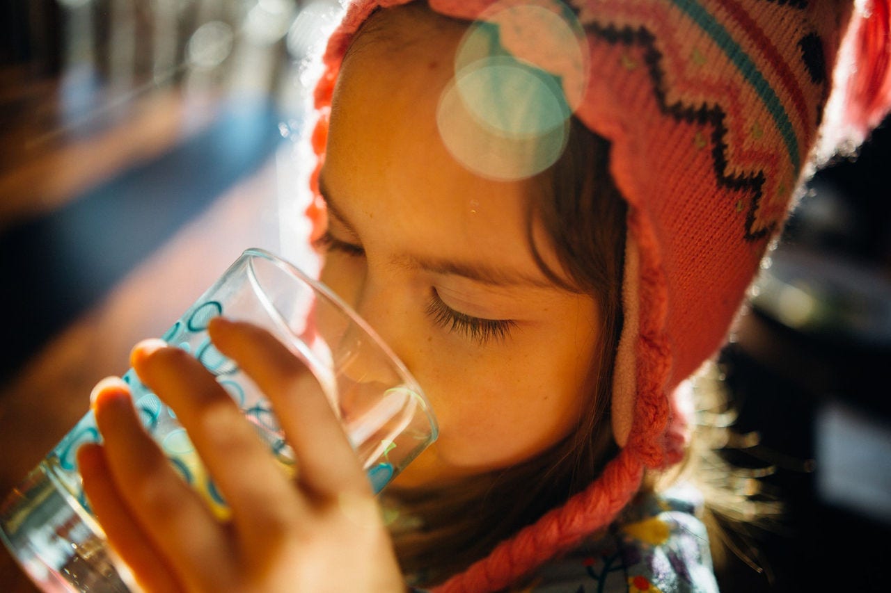 Niño con gorro tejido de lana bebiendo agua de un vaso de vidrio, con los ojos cerrados