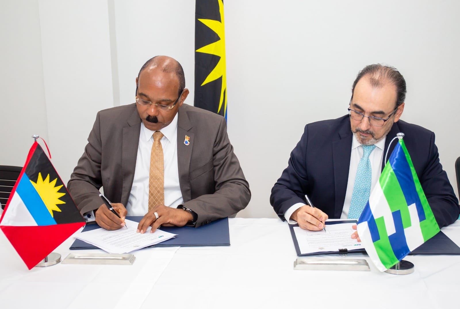 Dos autoridades firmando un acuerdo oficial junto a la bandera de Antigua y Barbuda y la bandera institucional.