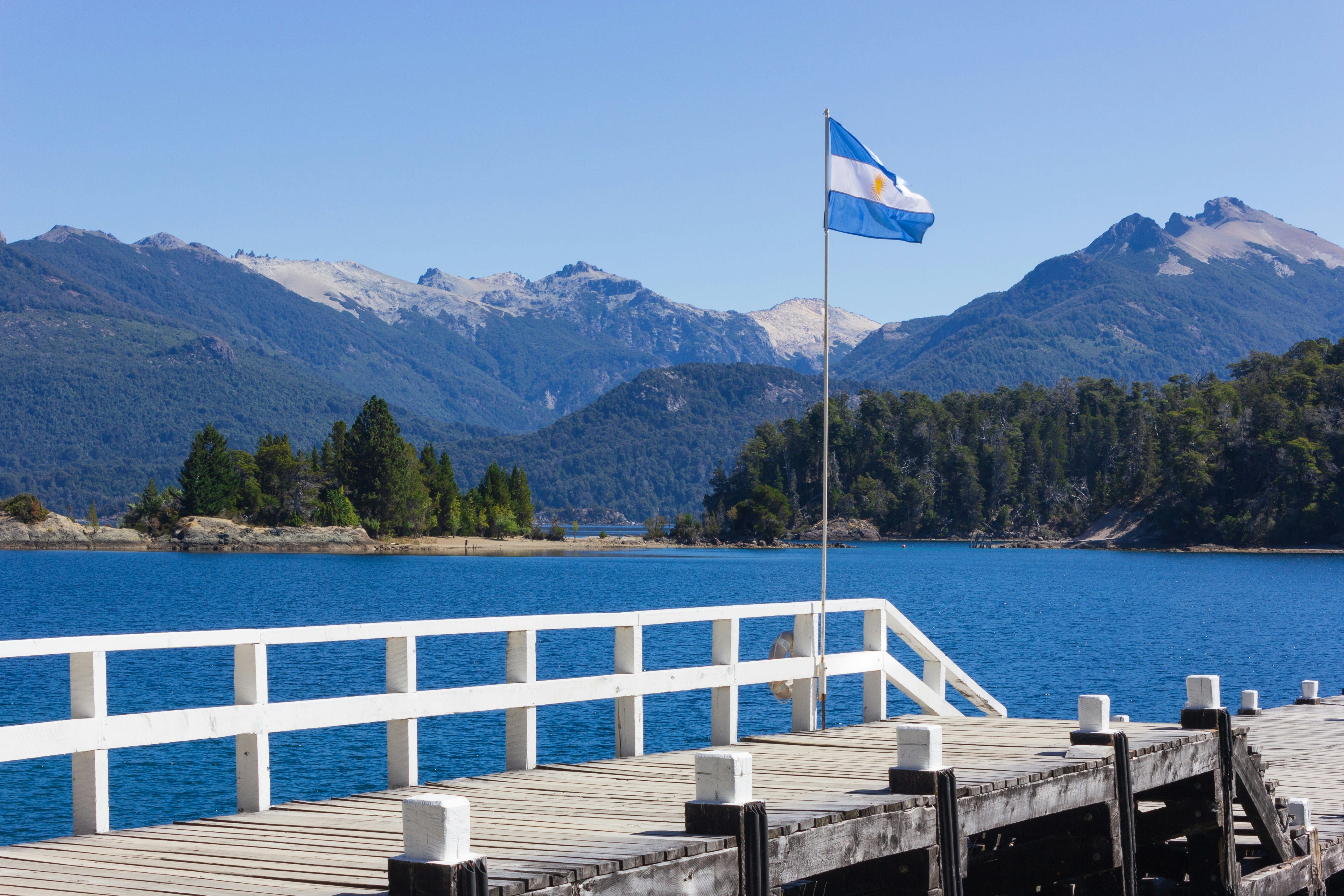 Muelle de madera frente a un lago andino con la bandera de Argentina ondeando ante las montañas.