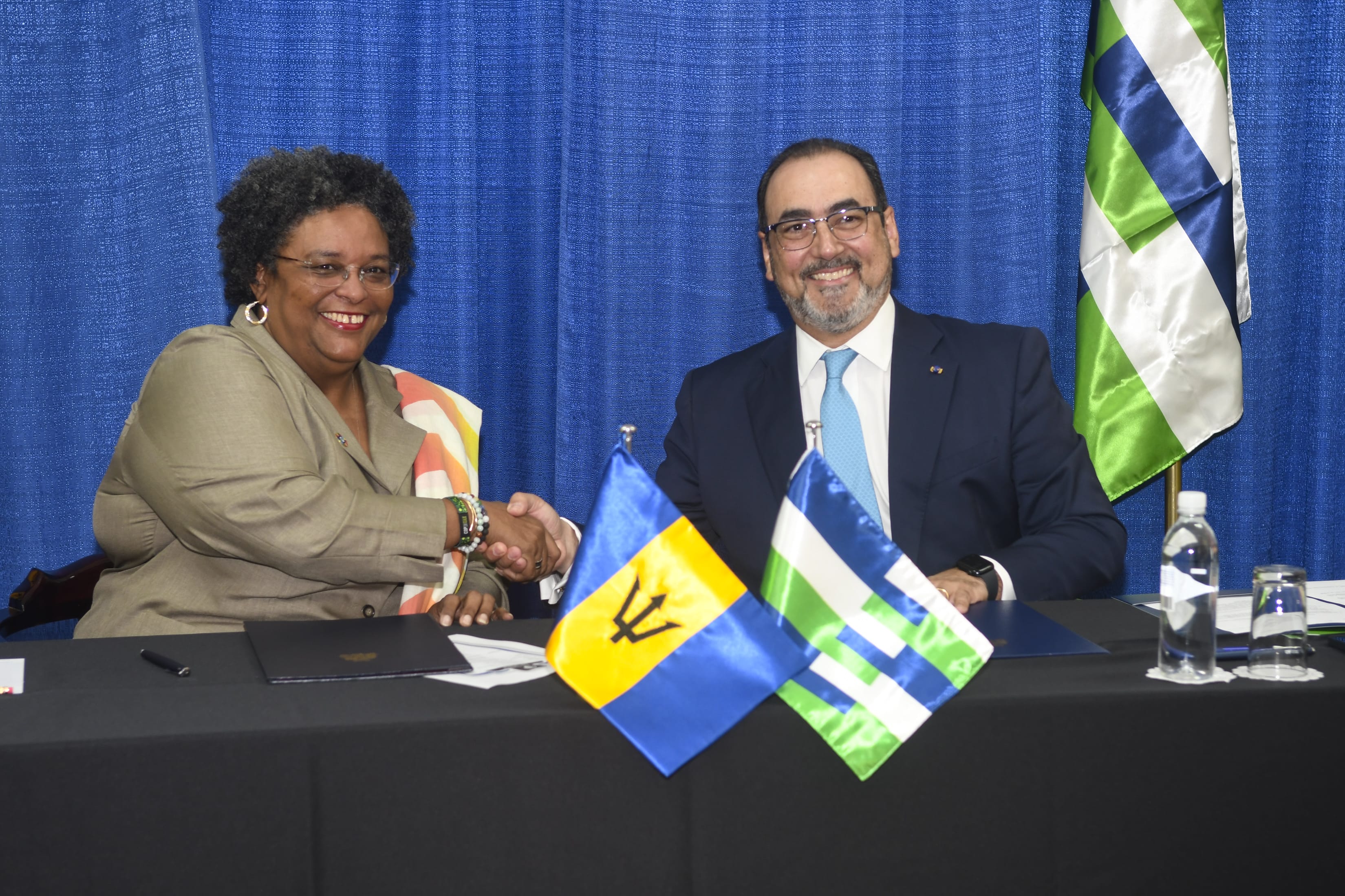 Representante institucional estrechando la mano de la Primera Ministra Mia Mottley junto a la bandera de Barbados.