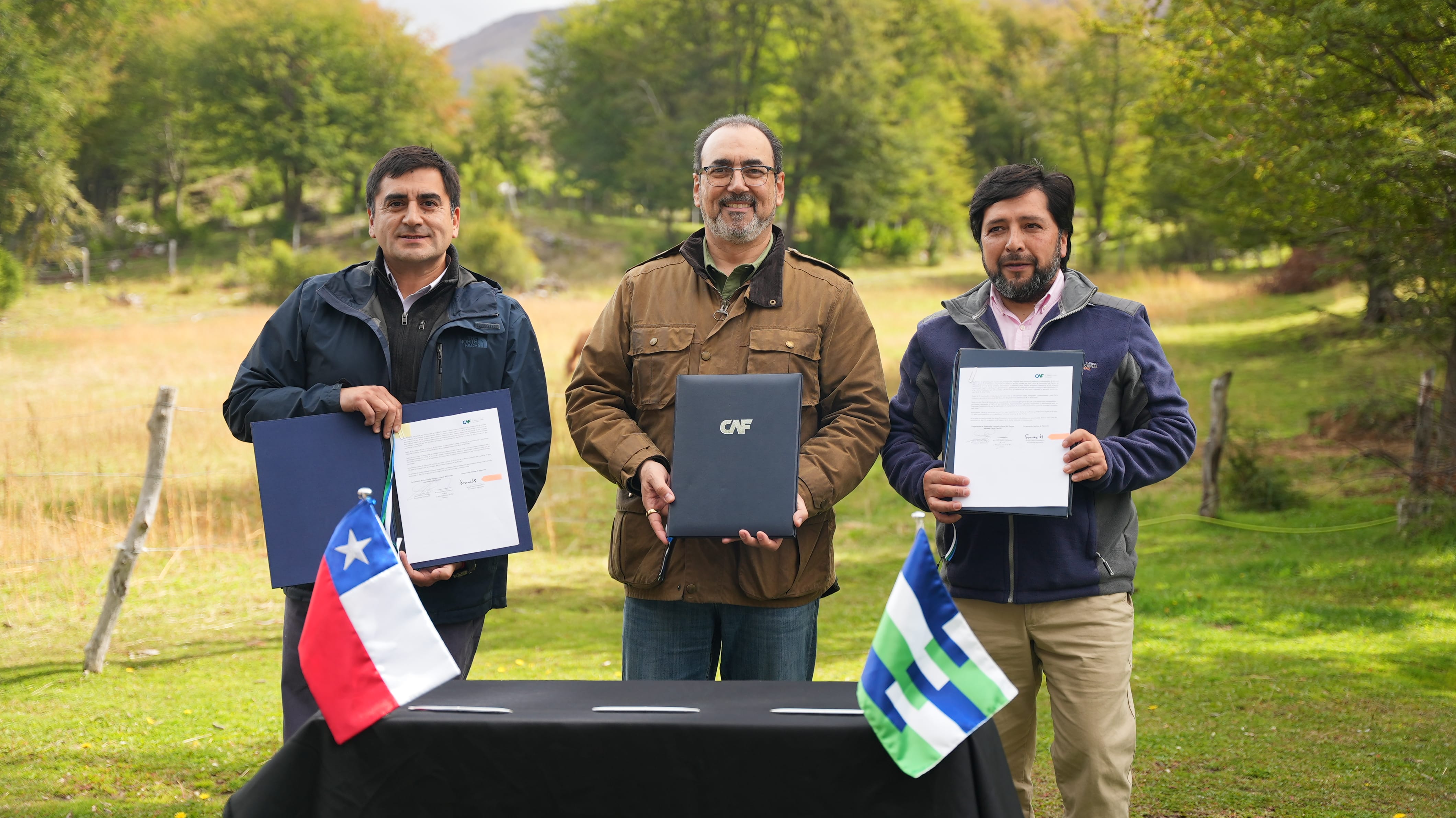 Tres representantes mostrando acuerdos firmados junto a las banderas de Chile en un entorno natural.