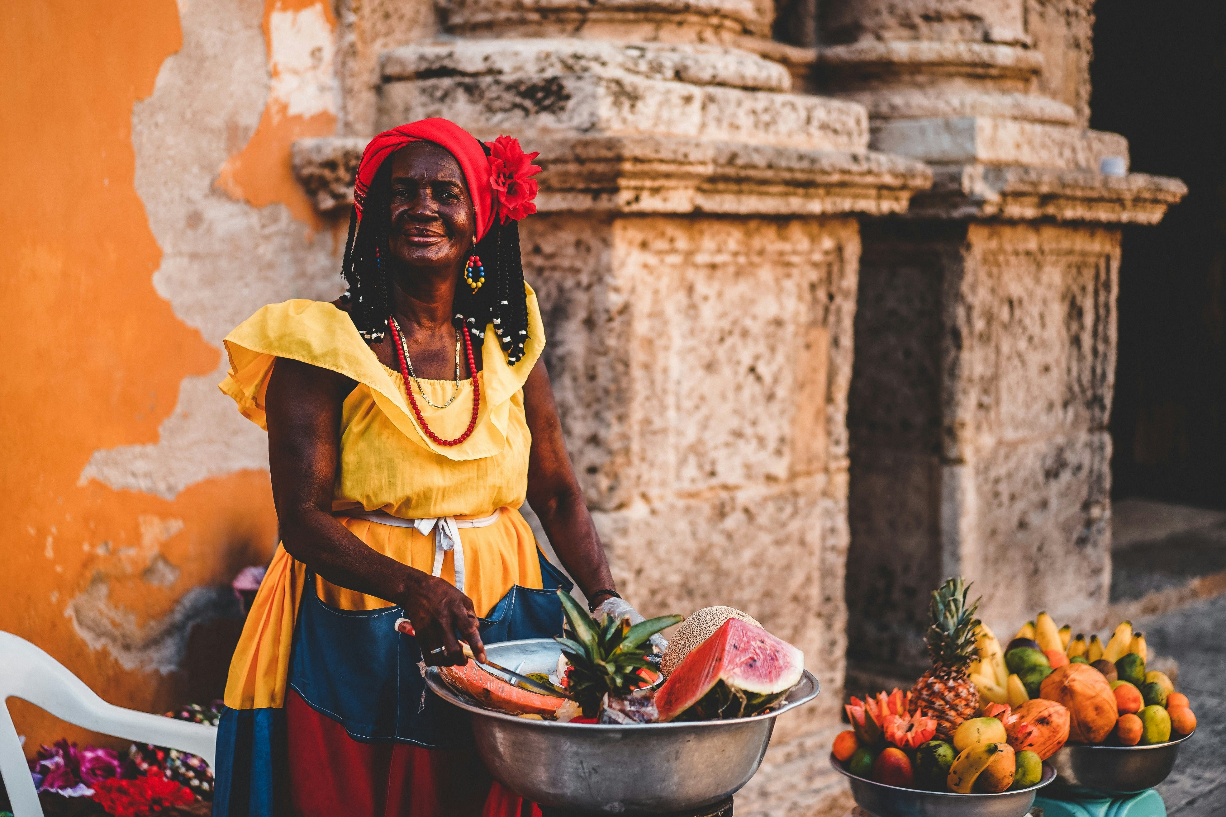 Palenquera en traje tradicional amarillo sentada con su ponchera de frutas frescas frente a un muro colonial.