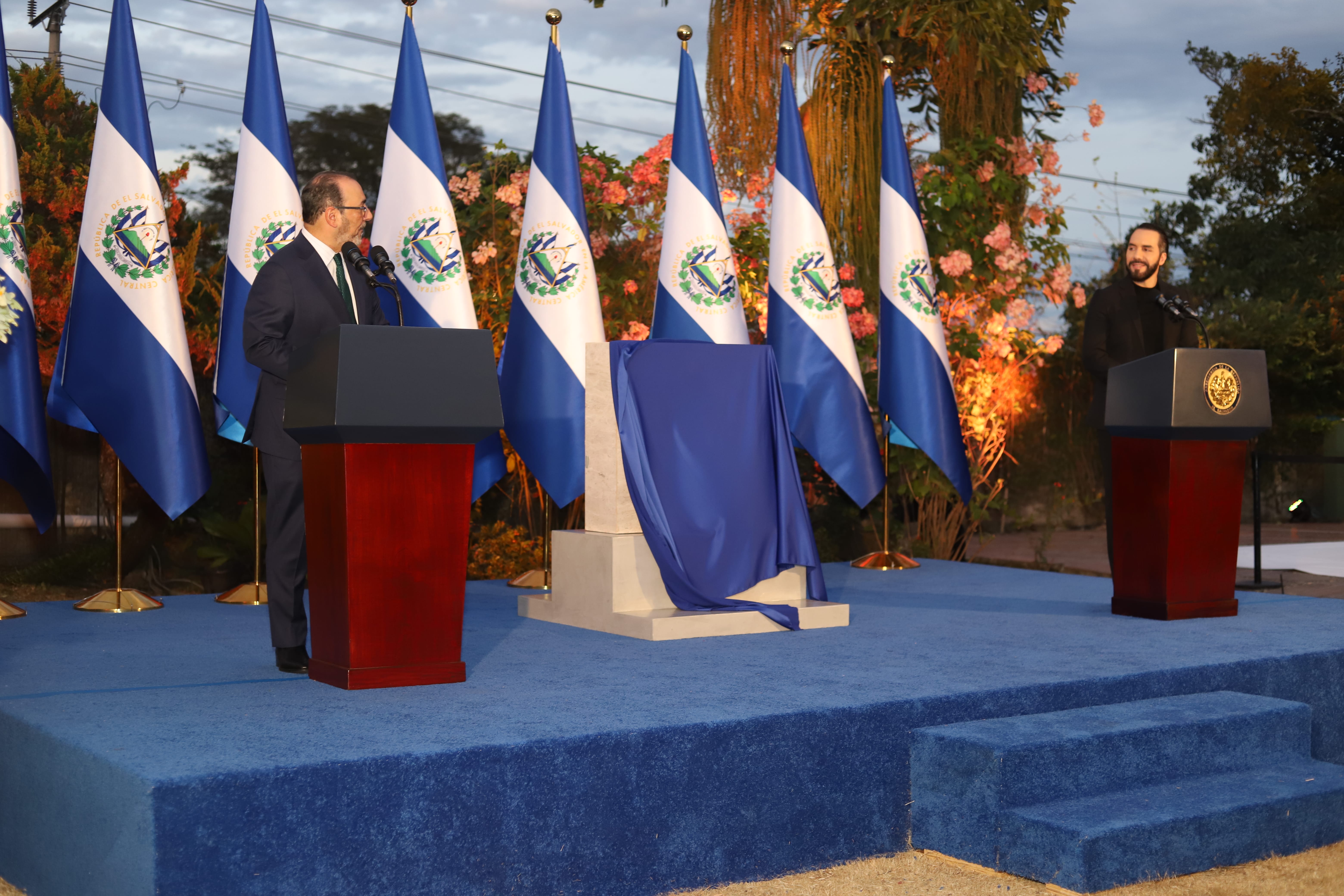 Autoridades en podio durante un evento gubernamental ante filas de banderas de El Salvador.