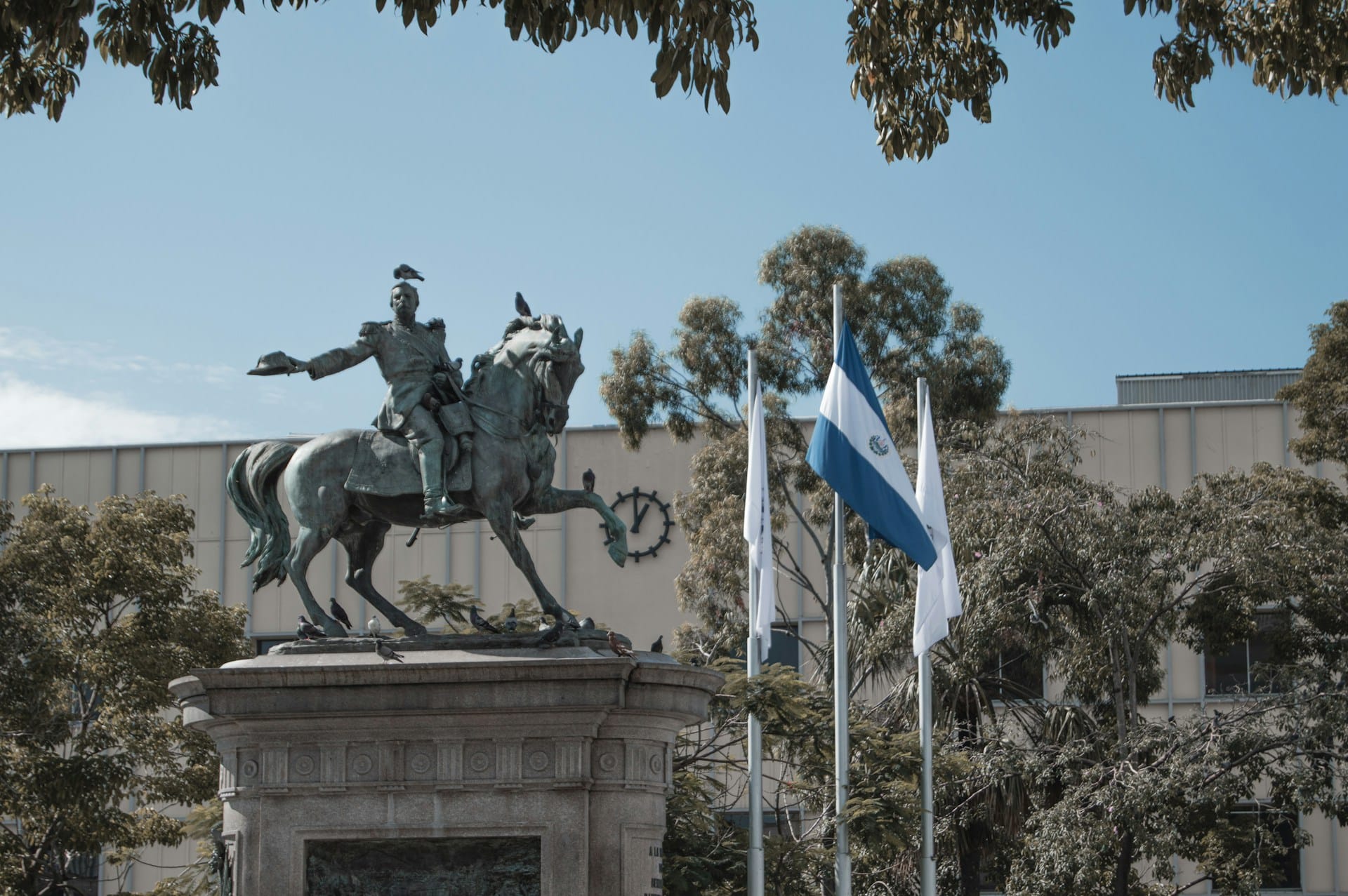 Estatua ecuestre de bronce sobre pedestal en la Plaza Gerardo Barrios con la bandera de El Salvador al fondo.