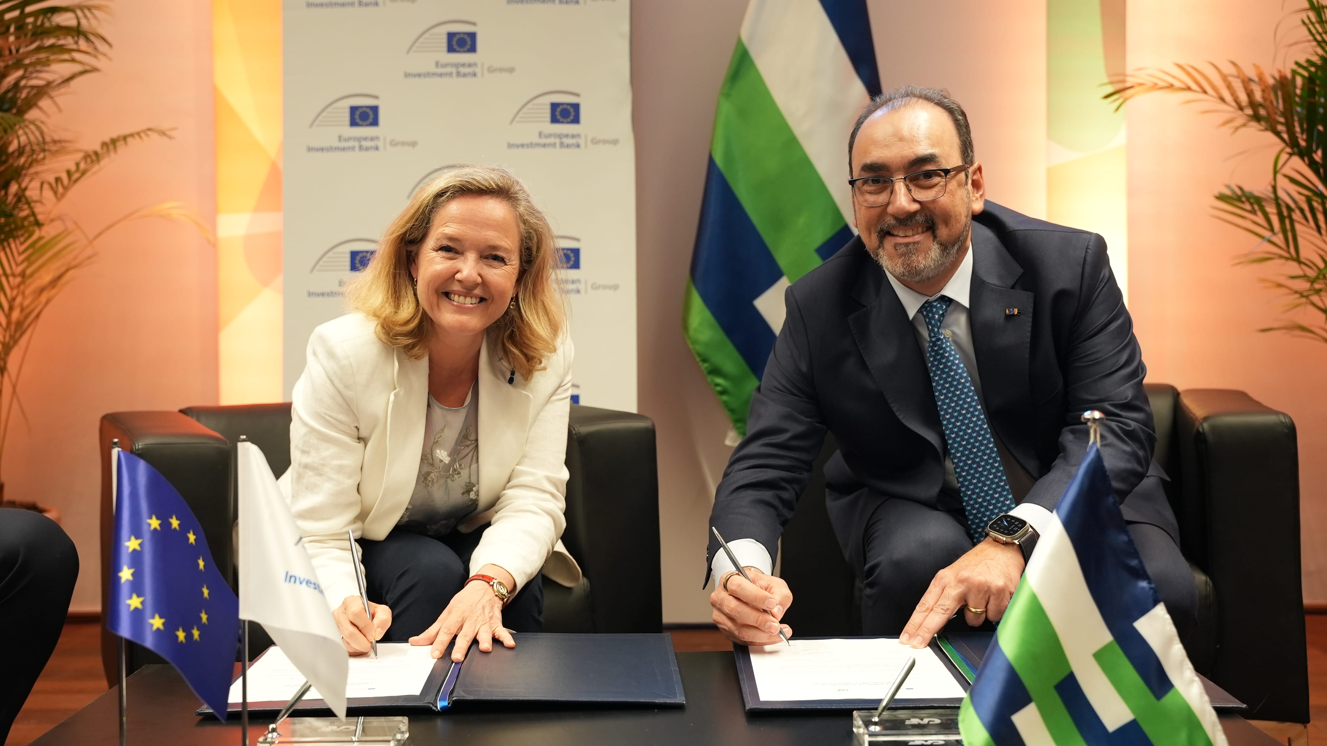 Dos autoridades firmando un acuerdo bilateral con la bandera de la Unión Europea y la bandera institucional al fondo.