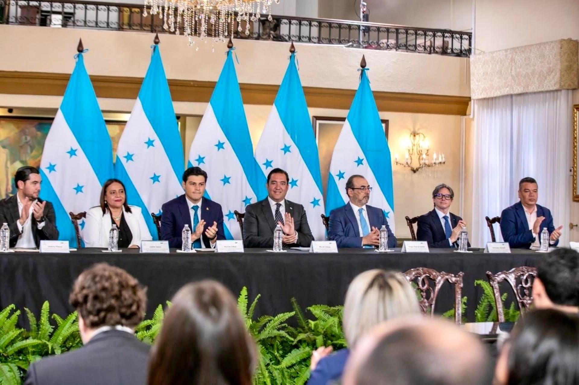 Representantes institucionales alineados en mesa directiva durante una conferencia con banderas de Honduras al fondo.