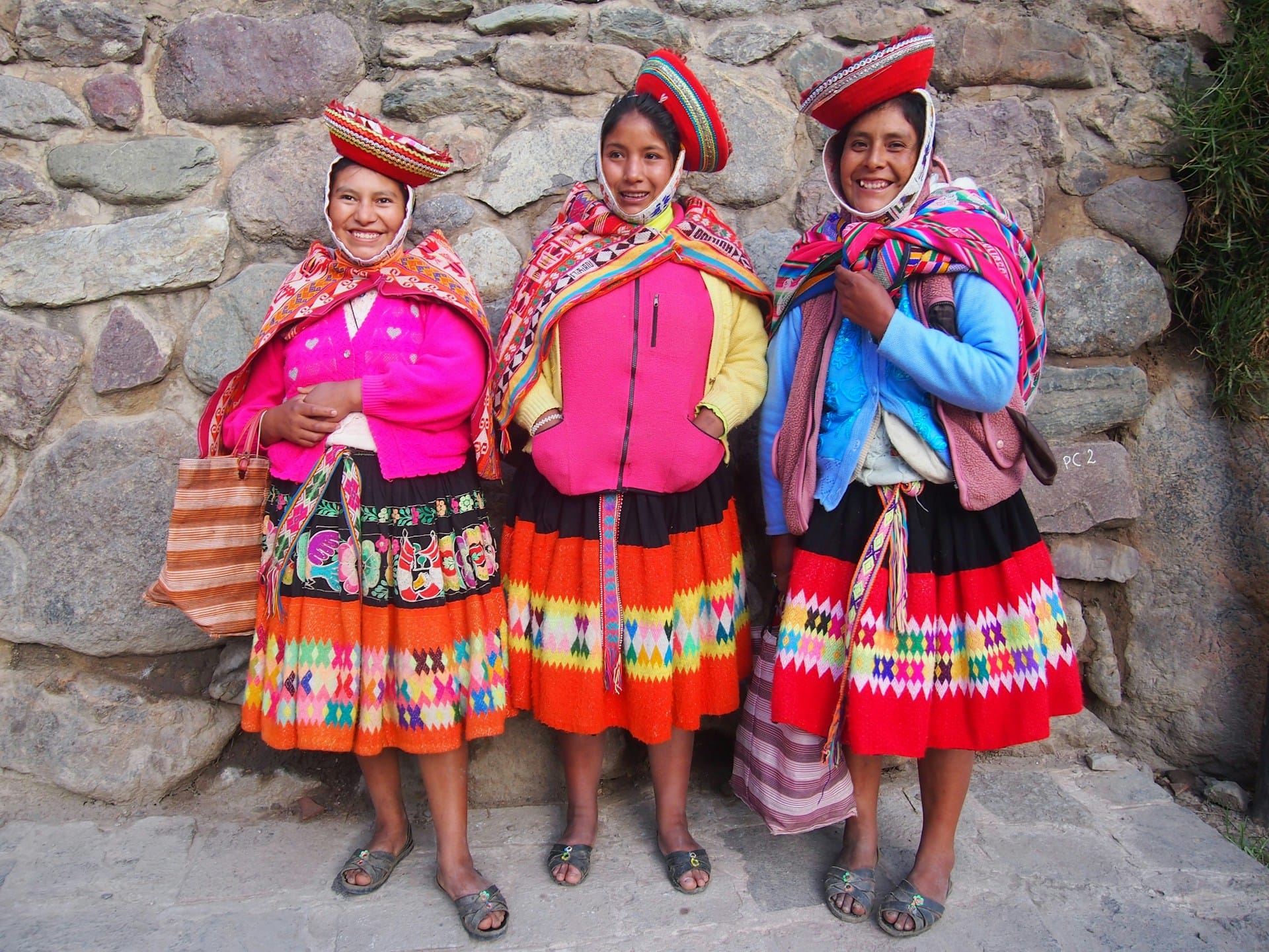 Tres mujeres peruanas con vestimenta típica andina y sombreros tradicionales junto a un muro de piedra.